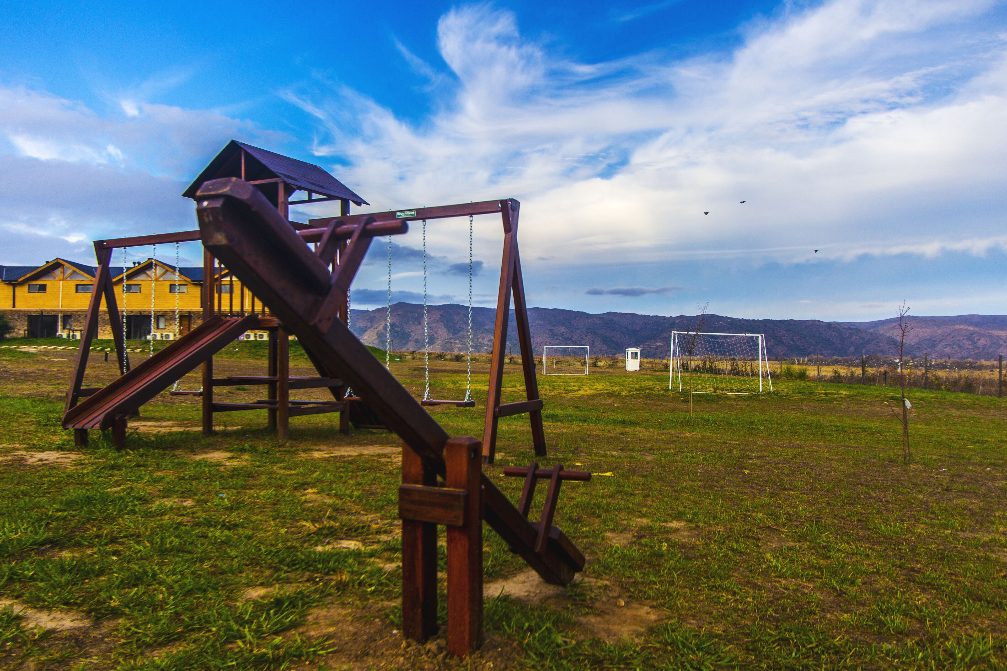 Ein Spielplatz mit Rutsche und Schaukeln, vor einer Berglandschaft und blauem Himmel.