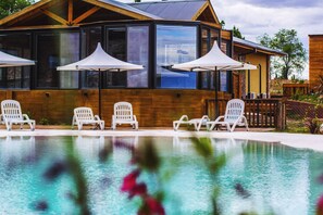 Un espace piscine avec des chaises longues blanches et des parasols, un bâtiment en bois doté de grandes fenêtres et un ciel dégagé.