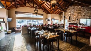 Un restaurante espacioso con vigas de madera y paredes de piedra, con varias mesas y sillas, ventanas amplias y una chimenea.