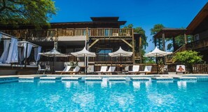 Un área de piscina con sillones de descanso y sombrillas, una terraza de madera con barandilla, y un edificio con varios balcones.