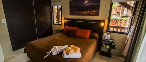 Quarto de hotel com uma cama grande, uma pintura do céu noturno, uma janela com vista para edifícios e uma mesa de cabeceira com uma luminária e uma mesinha.