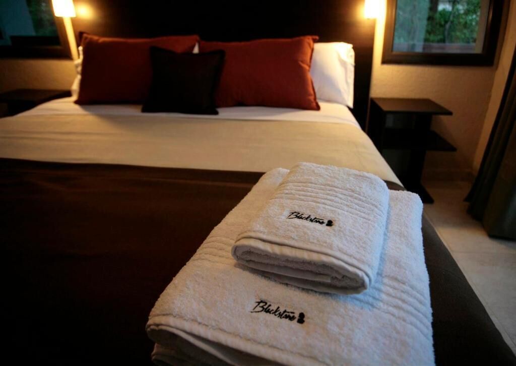 A neatly made bed with white linens and two pillows, a folded towel with the hotel name, and a nightstand with a lamp.