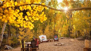 An outdoor seating area with Adirondack chairs and a wooden table, surrounded by trees with yellow leaves. - Mt. Harvard Chalet at Creekside chalets (Salida)