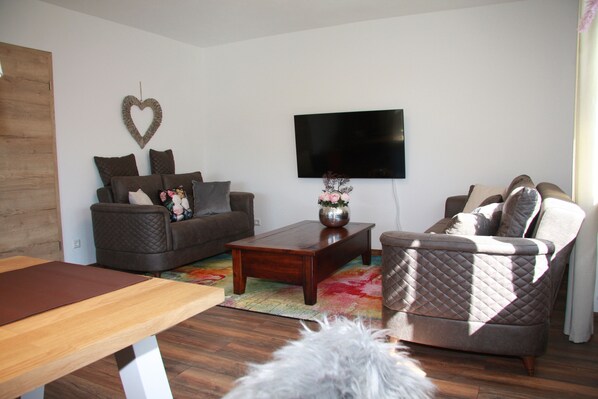 A living room with a brown sofa, a wooden coffee table, and a grey armchair. A flat-screen TV is mounted on the wall.