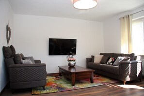 A living room with a dark brown sofa, a matching armchair, a wooden coffee table, and a flat-screen TV mounted on the wall.