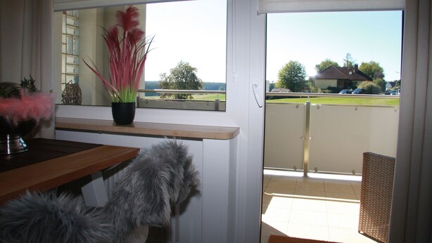 A room with a view of a green landscape, a window with blinds, a wooden table, and a wicker chair. - Fichtelherz Herzerl (barrier-free) - 89 m² - 2 bedrooms - balcony - max. 5 pers. (Thierstein)
