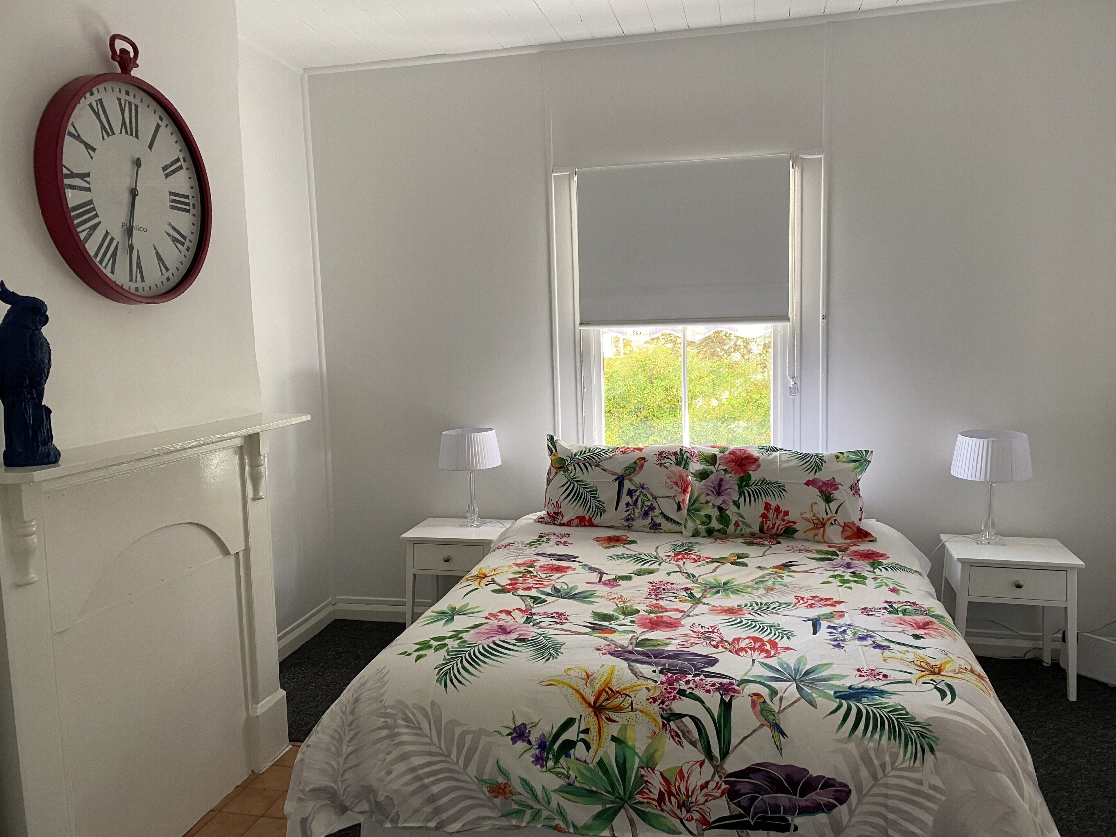 Ein Schlafzimmer mit floralem Bettbezug, einer Wanduhr, einem Fenster mit Blick ins Grüne und einem weißen Kamin.
