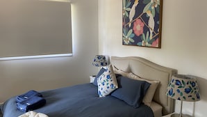A bedroom with a bed, two bedside lamps, a window with a blind, and a floral artwork on the wall.