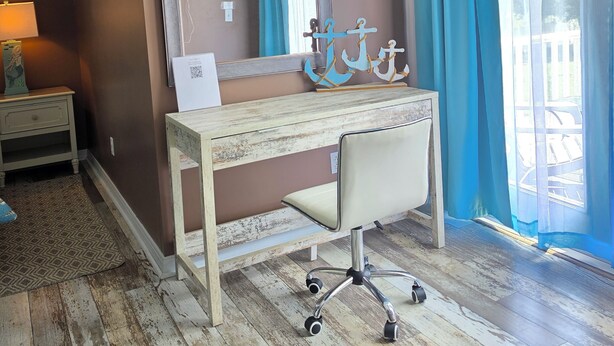 A room with a wooden desk, a large mirror, a 'Welcome' sign, a small statue, and a view through a window with blue curtains. - Mermaid relaxing studio with Pool (St. Augustine)