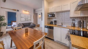 A compact kitchen with white cabinets, a microwave, and a dishwasher. There is a wooden dining table with chairs, a sofa, and a small plant. - 2-Bed River View | Private Terrace+Sauna (Quebec City)