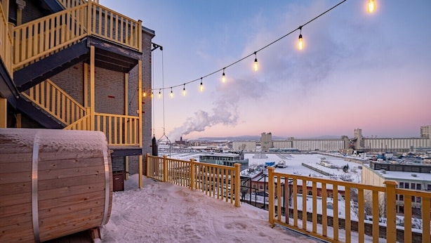A snowy outdoor area with a wooden barrel, a yellow railing, and a view of a cityscape with buildings and a bridge in the background. - 2-Bed River View | Private Terrace+Sauna (Quebec City)