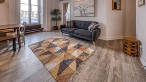 A living room with a dark grey sofa, a wooden dining table with chairs, a patterned rug, and a large window with a view of a snowy deck. - 2-Bed River View | Private Terrace+Sauna (Quebec City)