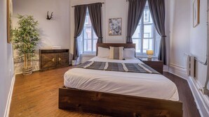 A bedroom with a wooden bed, two windows with curtains, a bedside table with a lamp, a wall-mounted artwork, and a potted plant. - 2-Bed River View | Private Terrace+Sauna (Quebec City)