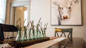 A dining table with a wooden tray holding several glass vases with dried plants. A framed artwork of a cow is on the wall behind the table. - Circle Bar Manor — Spacious 4BR, Sheridan WY (Sheridan)