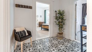 Een hal met vloertegels met patroon, een stoel met kussen, een kamerplant en een deur naar een andere kamer.