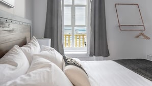 Een netjes opgemaakt bed met witte lakens, een houten hoofdbord en uitzicht op een balkon met gele leuningen.