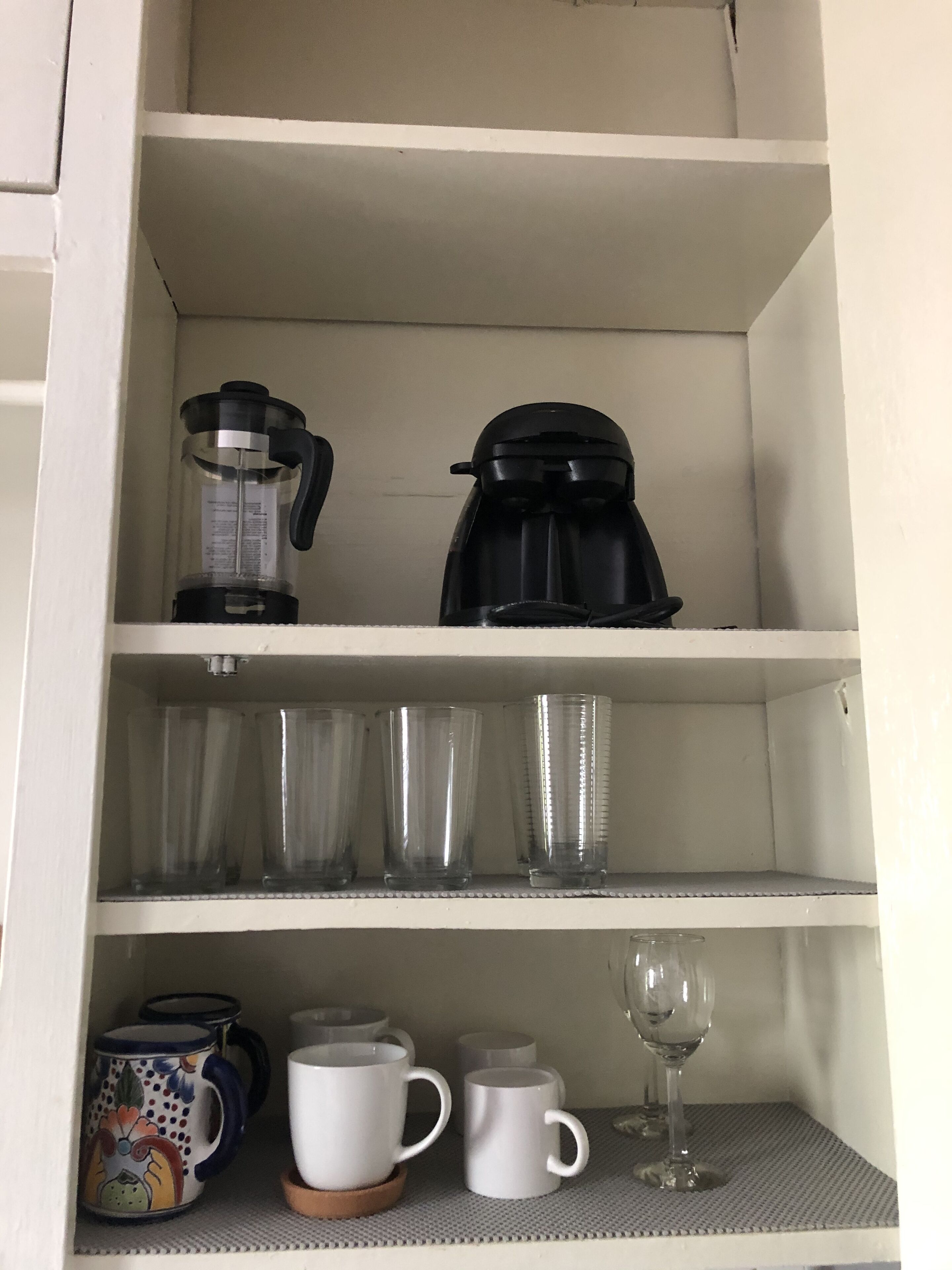 Una repisa de cocina con una cafetera, vasos transparentes y tazas blancas.