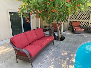 A wicker sofa with red cushions, a tree with orange flowers, a pool, and a wooden fence. - Hidden Oasis with pool and garden (Fair Oaks)