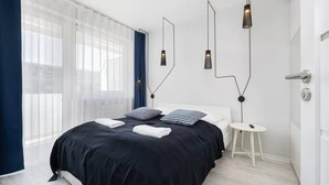 A modern bedroom with a large bed, a small white side table, and a window with sheer curtains. - Seaside Apartment | 400 m to the Beach, Gym, Balcony (Kołobrzeg)