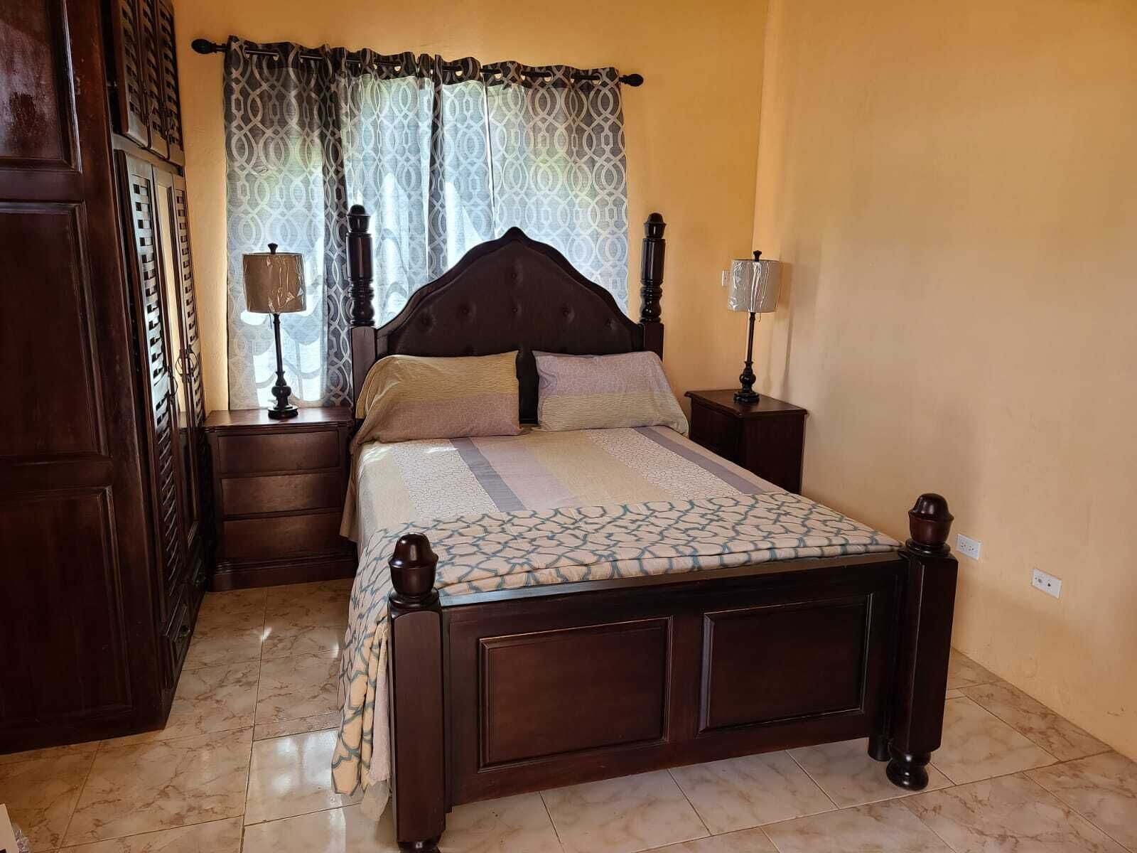 Une chambre à coucher avec un lit en bois, deux tables de chevet, une commode, une couvre-lit à motifs et une fenêtre ornée de rideaux à motifs.