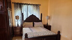 Una camera da letto con un letto di legno, due comodini, un tavolino e una coperta con motivi decorativi, oltre a una finestra con tende a fantasia.