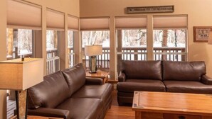 Ein geräumiges Wohnzimmer mit einem großen braunen Ledersofa, einem Holztisch und einem Deckenventilator. Große Fenster bieten Blick auf Bäume, und ein Schild mit der Aufschrift "Welcome to the Mountain".