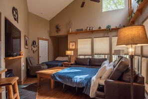 A cozy living room with a large bed, a sofa, a coffee table, and a TV. There are two windows with blinds, a ceiling fan, and a shelf with decorative items.