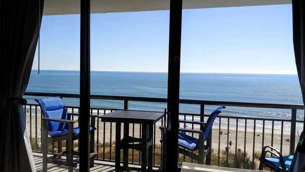 A balcony with a table and blue chairs, overlooking a beach and the ocean. - Beautiful Clean Modern Direct Oceanfront Unit With Amazing Views (Myrtle Beach)