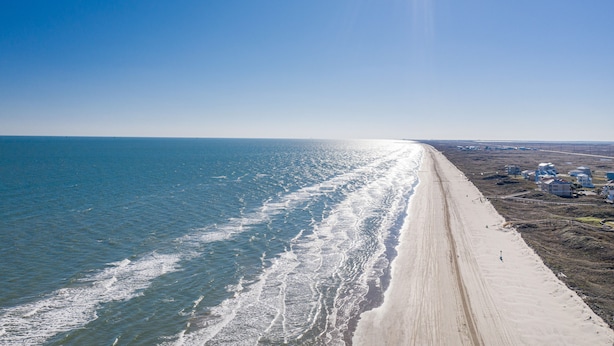 Uma praia com uma extensa faixa de areia, céu azul límpido e alguns edifícios ao longe.