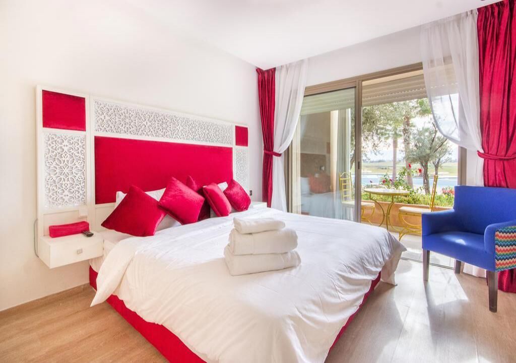 A hotel room with a large bed, red and white decor, a blue chair, and a view of the outdoors.
