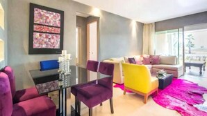 Een moderne woonkamer met een glazen tafel, paarse en gele stoelen, een bank en een groot raam.