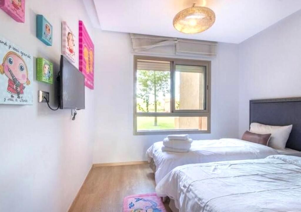 A bedroom with two beds, a window with a view of greenery, a ceiling light fixture, and wall decorations.