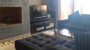 En stue med et fladskærms-tv på et sort stativ, et sofabord i glas og en mørk sofa.