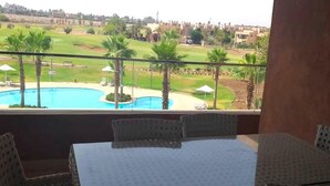 En balkon med et bord og stole, med udsigt over en swimmingpool og en golfbane.