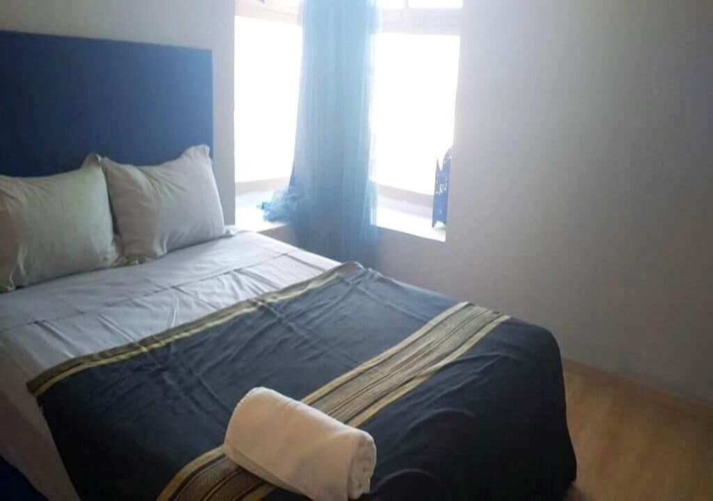 A neatly made bed with a blue headboard, a window with curtains, and a wooden floor.