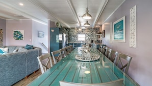 Comedor con mesa y sillas de vidrio, cocina al fondo y sala de estar con sofá y cojines decorativos.