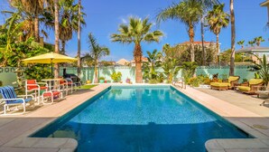 Una piscina con sillas de descanso y sombrillas, rodeada de palmeras y una valla decorativa.