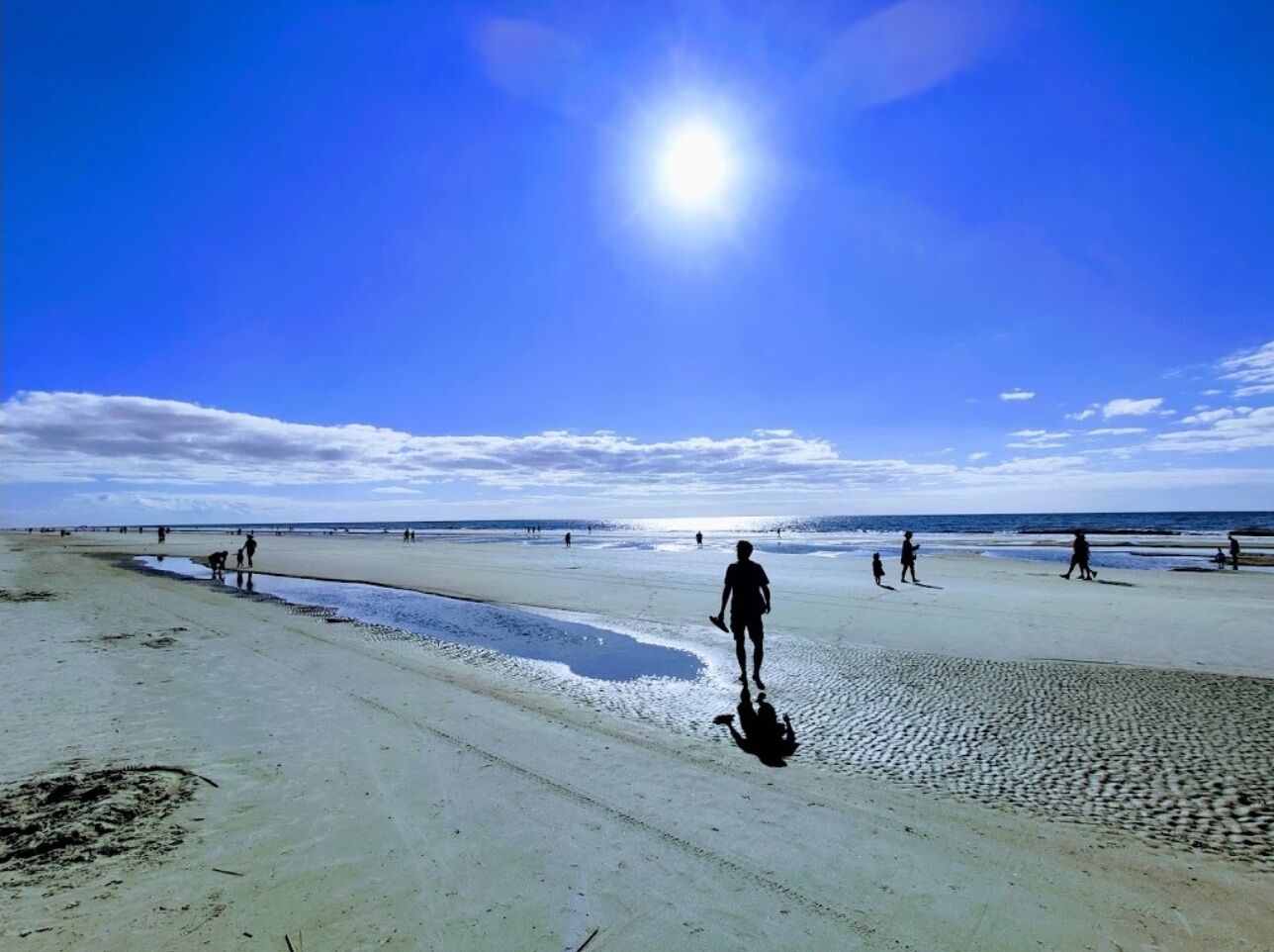 Una playa con gente caminando y parada, cielo despejado y el sol brillando.