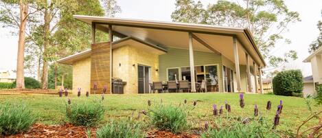 A modern house with a covered patio, a stone fireplace, and a well-maintained lawn with purple flowers.