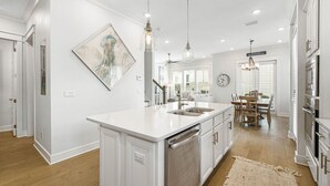 Une cuisine moderne avec des meubles blancs, un îlot central, des luminaires suspendus et un coin repas comprenant une table et des chaises en bois.