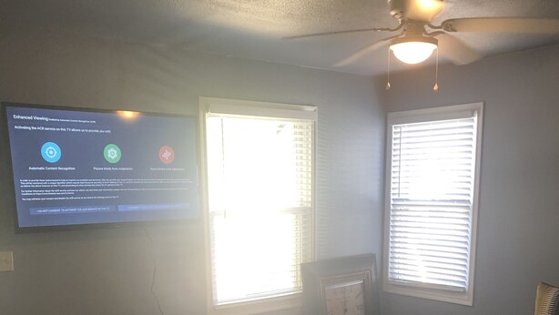Una sala de estar con un televisor de pantalla plana montado en la pared, dos ventanas con persianas, un ventilador de techo y suelo de madera.