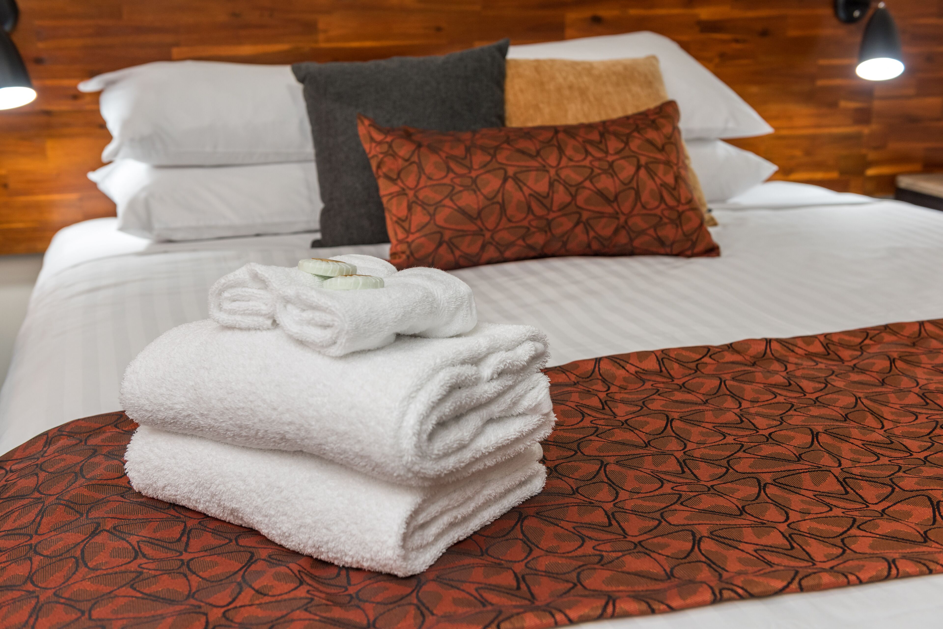 A neatly made bed with white linens, a red patterned throw, and a stack of white towels.