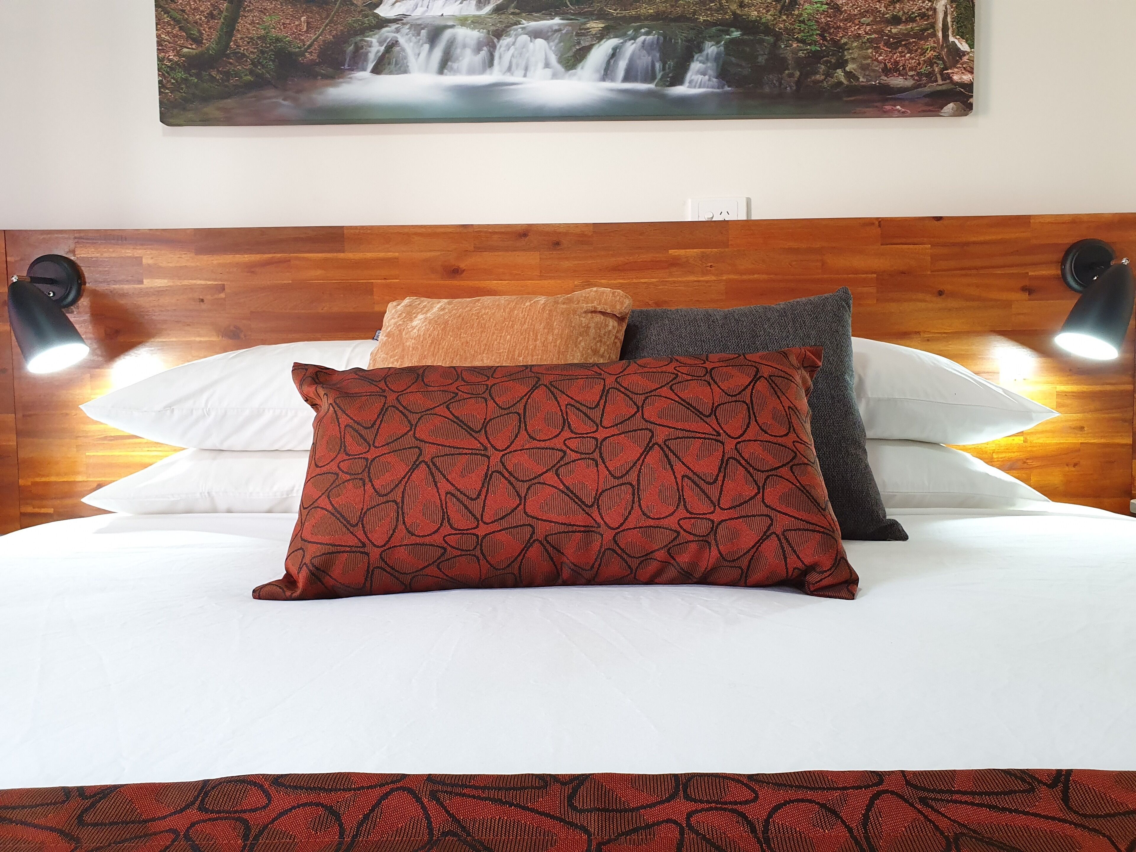 A neatly made bed with white and patterned pillows, a wooden headboard, and two wall-mounted reading lamps.