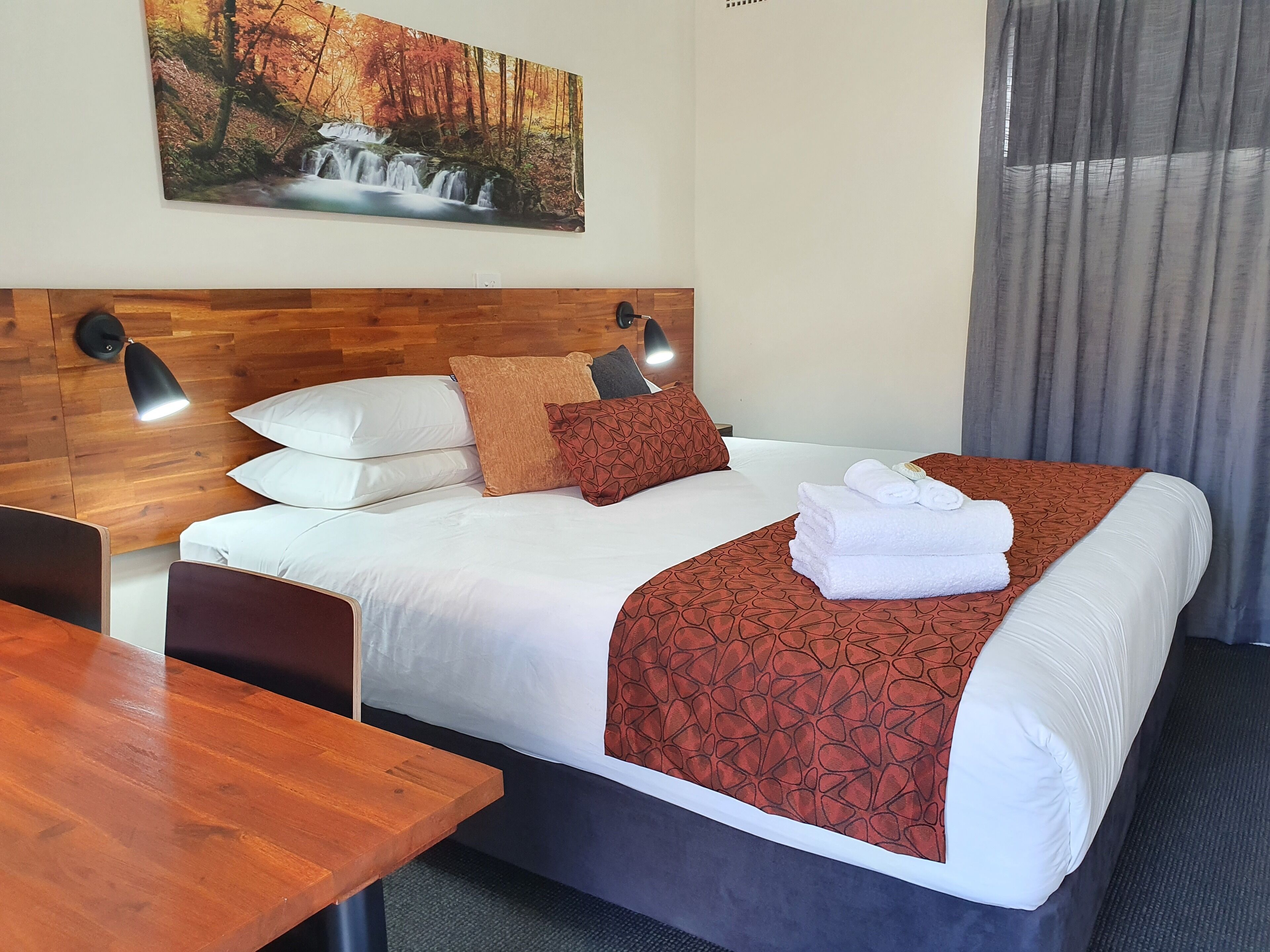 Habitación de hotel con cabecera de madera, cama con ropa blanca y una manta roja estampada, cama bien hecha con toallas, una mesa de madera y un cuadro de una cascada en la pared.