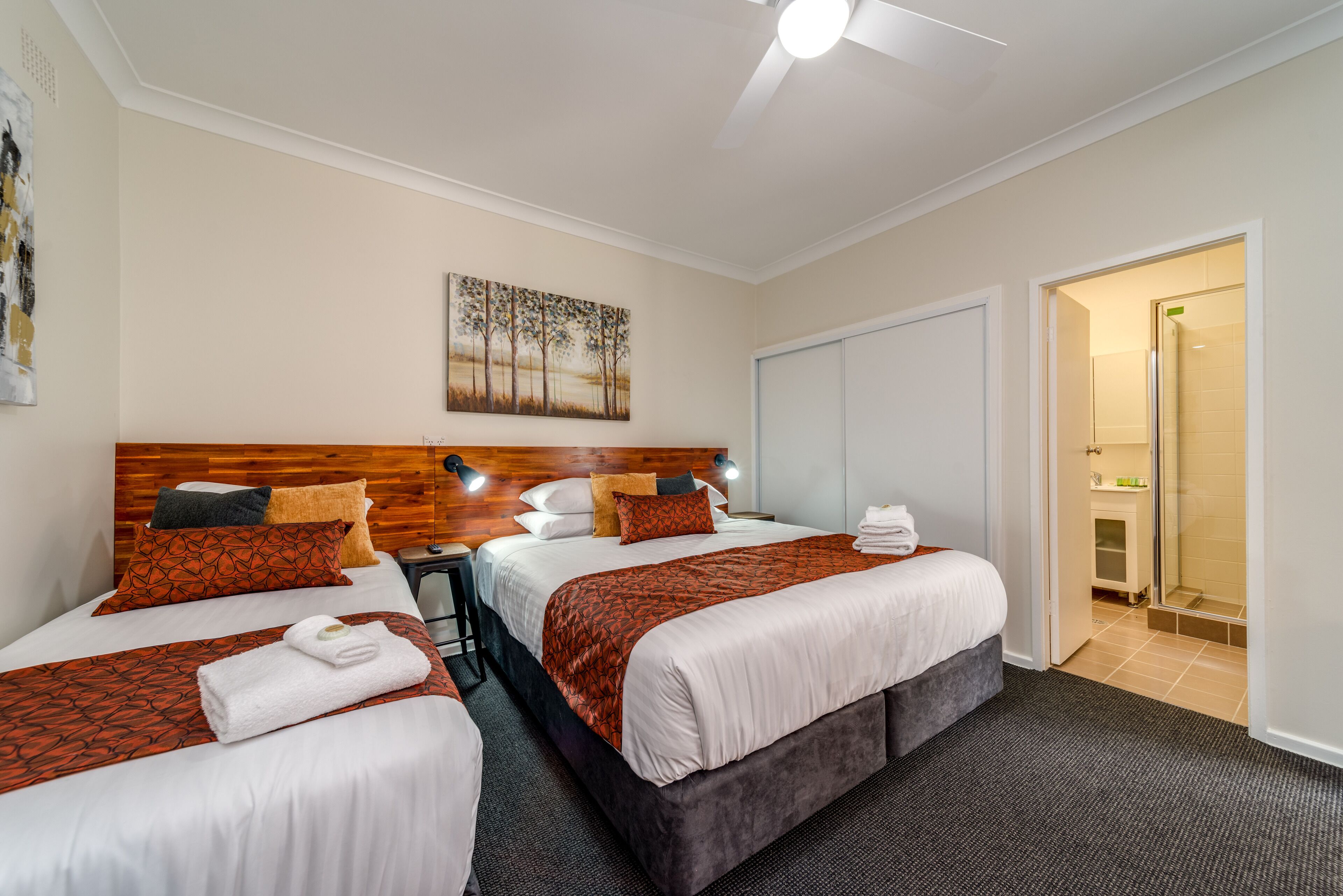 Une chambre d’hôtel avec deux lits, une tête de lit en bois, un tableau accroché au mur et un espace douche visible à travers une porte ouverte.