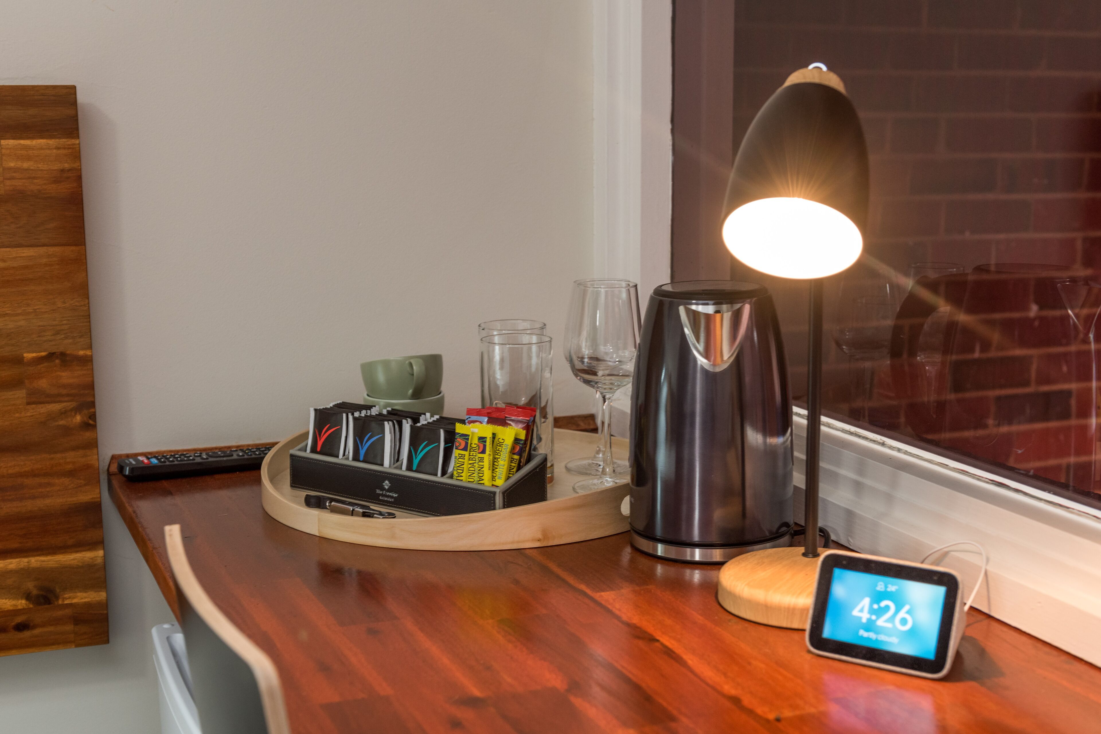 Una mesa de madera con una bandeja que contiene un control remoto, un vaso, una cafetera y un teléfono inteligente mostrando la hora.