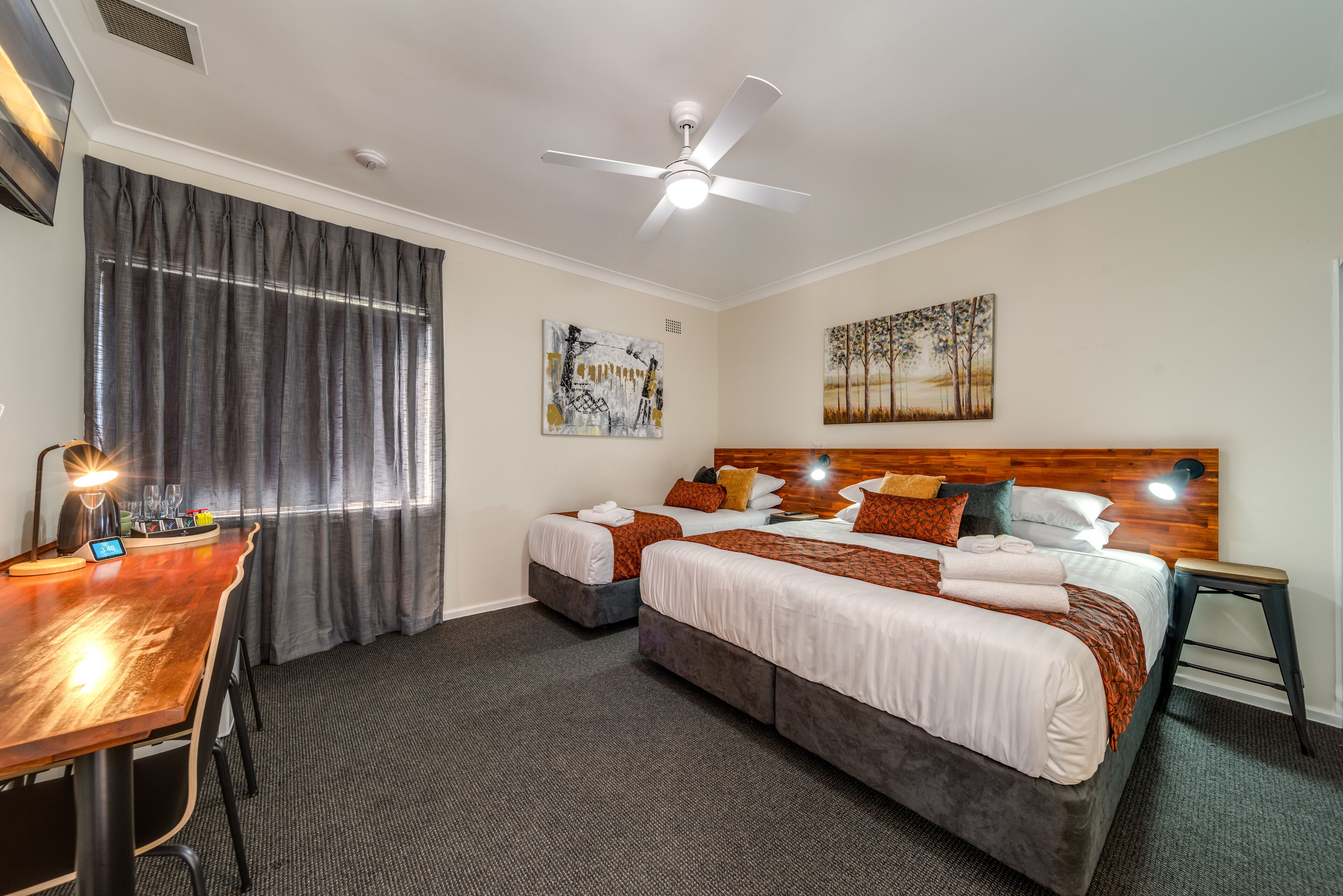 A hotel room with two beds, a desk, a ceiling fan, and artwork on the walls.