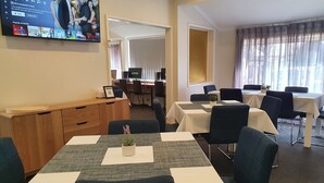 Quarto de hotel com televisão exibindo um programa, uma mesa com um laptop e um espaço de refeições com mesas e cadeiras.