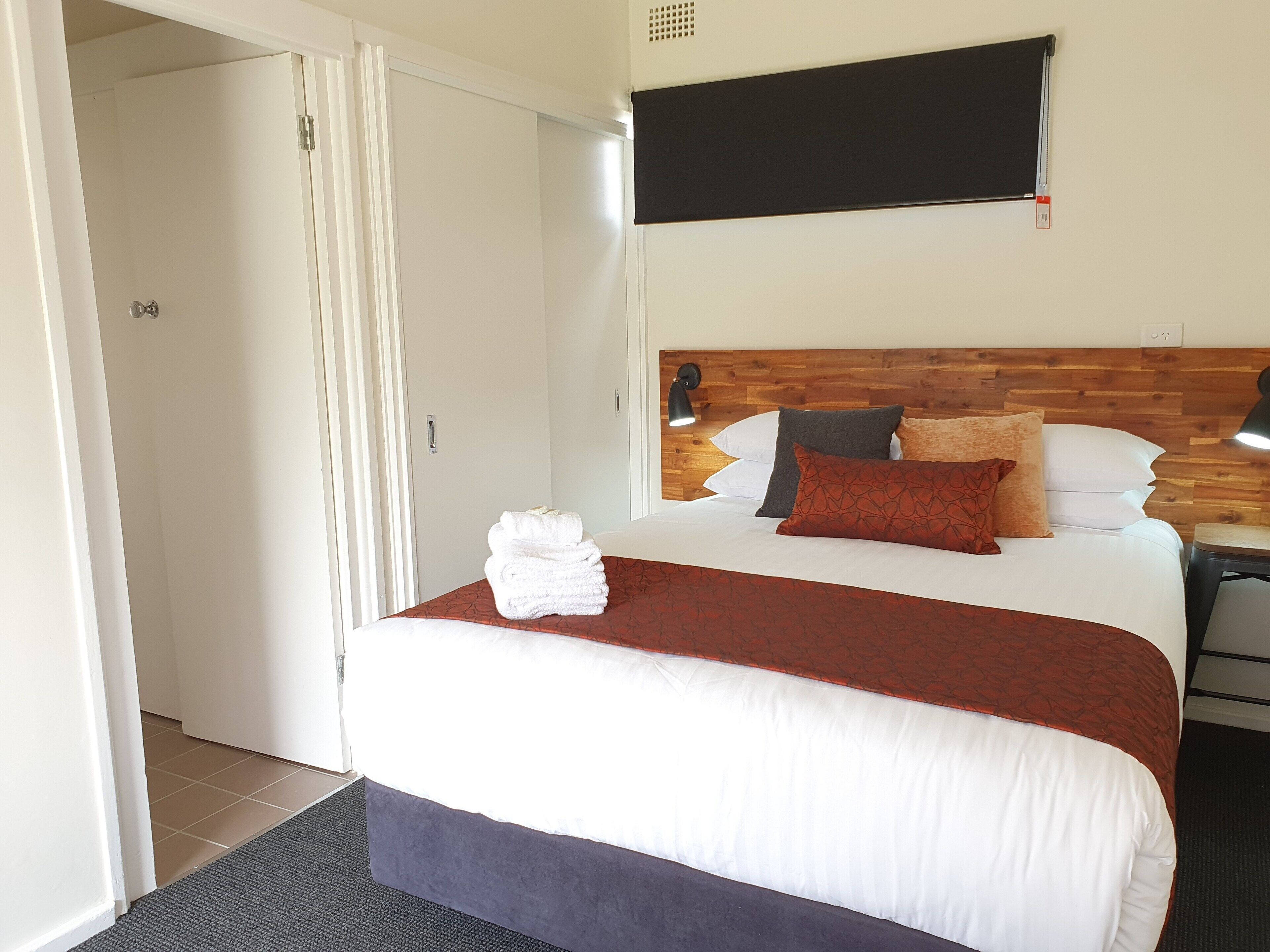 A hotel room with a neatly made bed, a wooden headboard, and a flat-screen TV mounted on the wall.