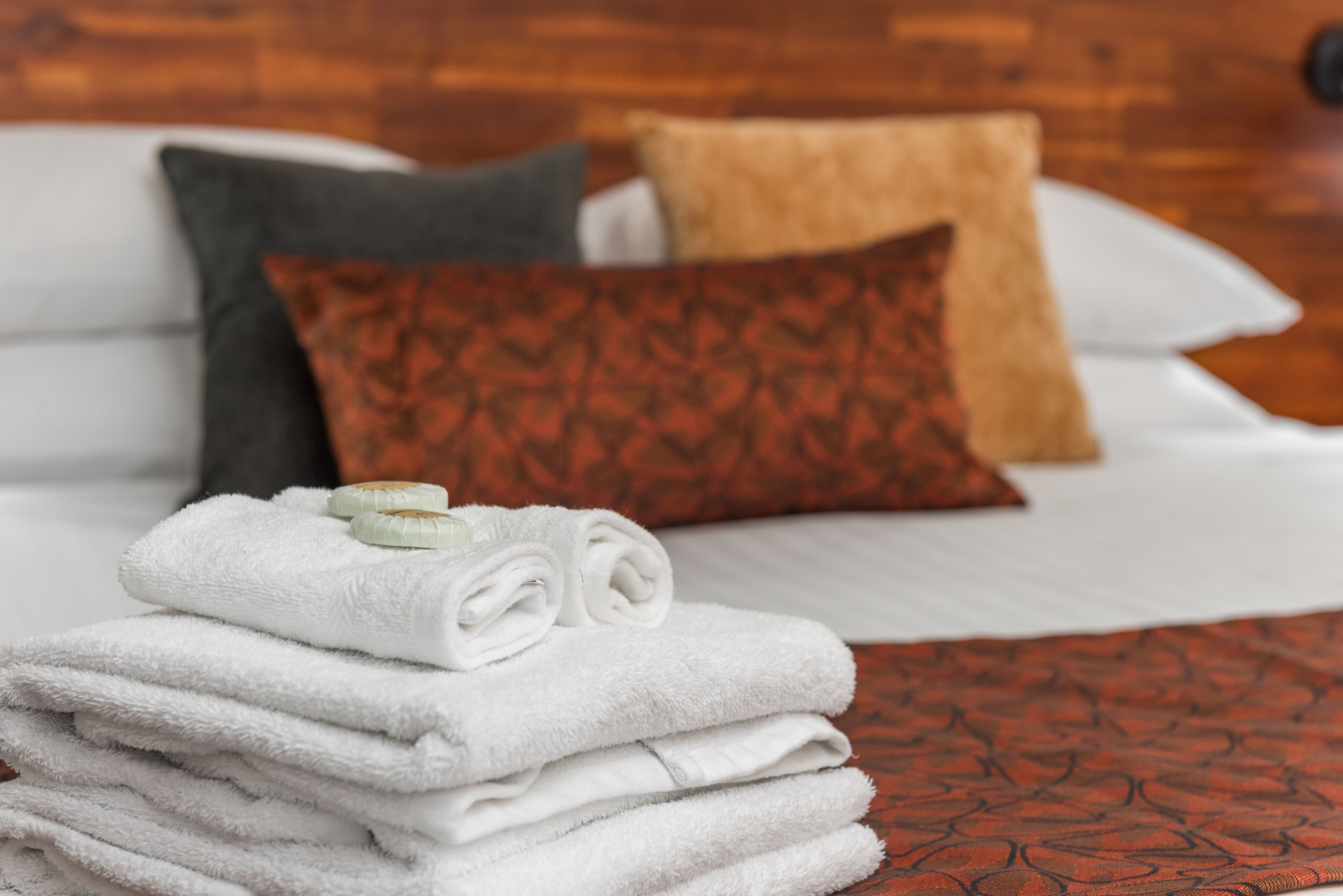 A neatly made bed with white bedding, a red patterned throw, and a stack of white towels.